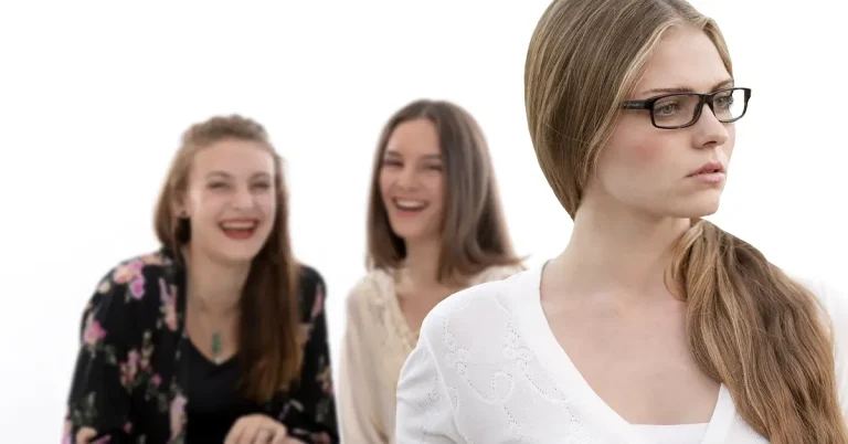 A sad young woman stands in the foreground whilst two other women are laughing at her behind her back.