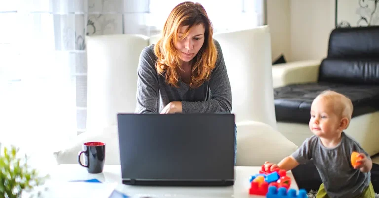 A woman is working at her laptop on a coffee table while her toddler son plays nearby with some building bricks.