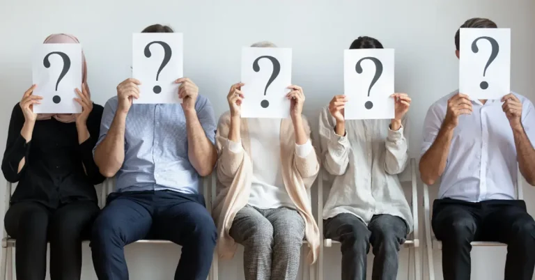 A line of job applicants sitting on chairs holding up question marks over their faces.