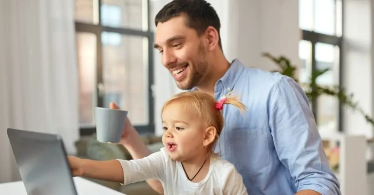 A dad and his young daughter are sitting at a table in their home in front of a laptop computer.