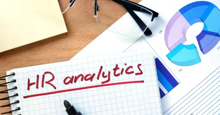 A notebook on a cluttered desk has the words 'HR Analytics' written on it.
