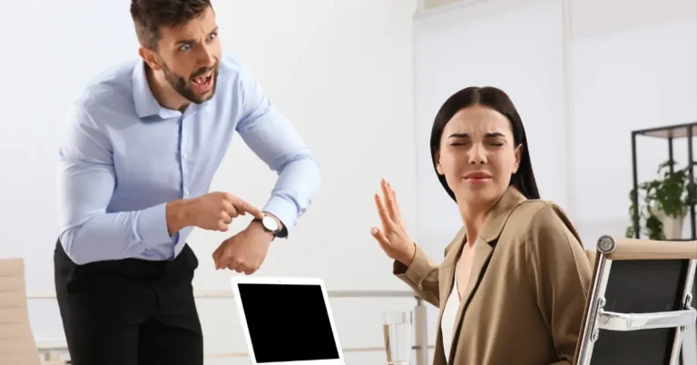 In an office, an angry boss is pointing to his watch whilst a female office worker is turning away.